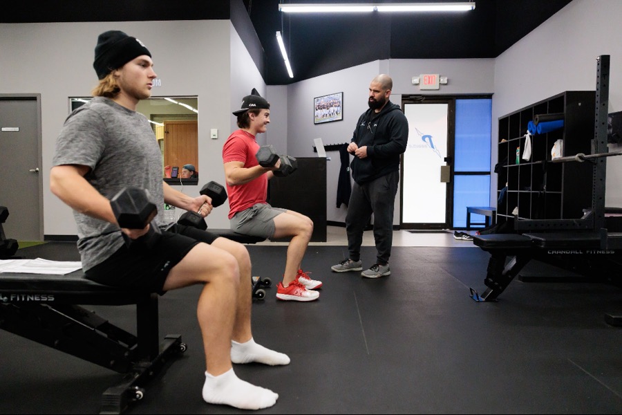Two baseball players doing seated dumbbell arm work while Jason Aguiar coaches