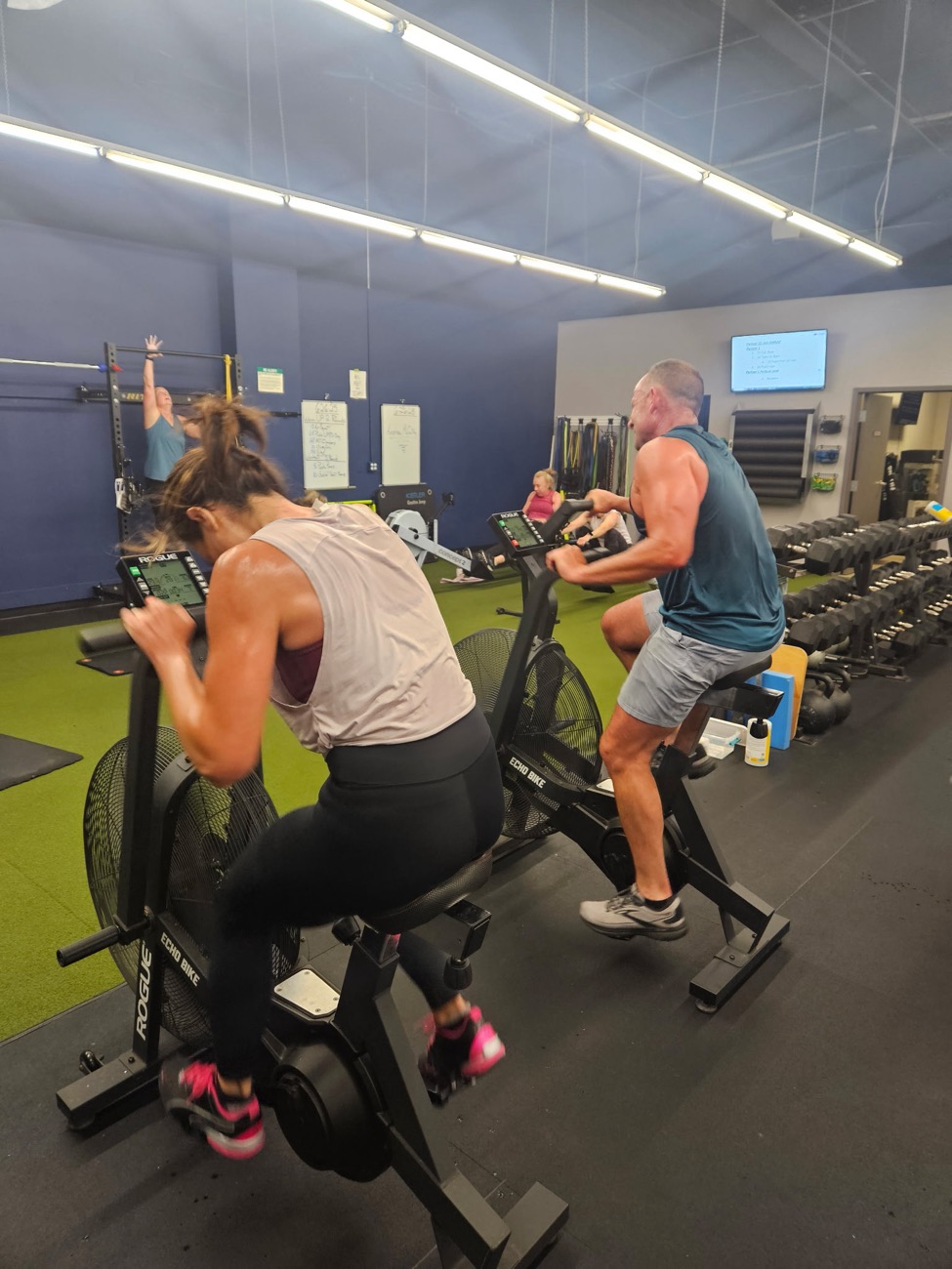 Adults in a Pinnacle group class riding Echo bikes while others row and train in the background